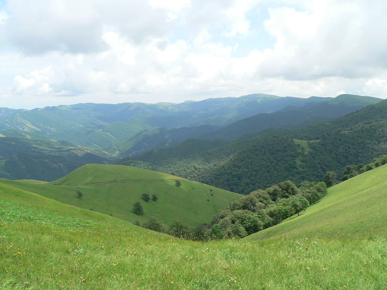 Wide view of lush green valleys and rolling hills with forested mountains in the distance under a partly cloudy sky