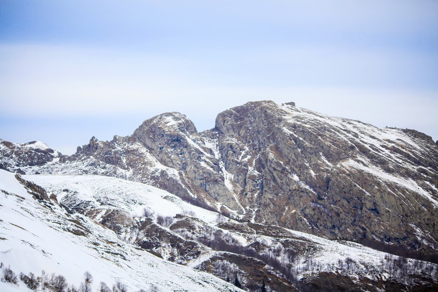 Snow-covered rocky mountain peaks with patches of snow on slopes, under a light blue sky with faint clouds