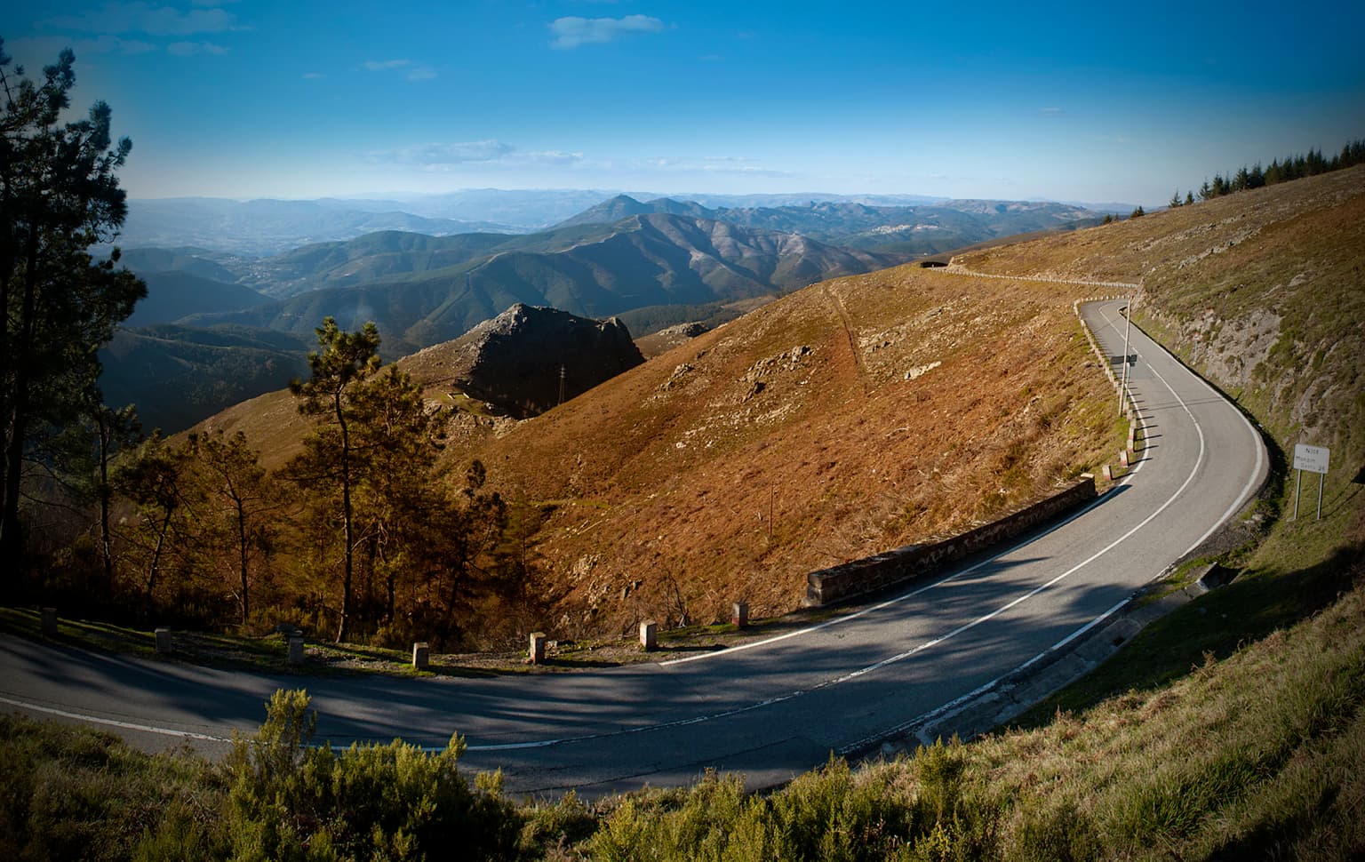 Winding paved road through mountainous landscape with dry brown vegetation, scattered trees, and distant mountain ranges under clear blue sky