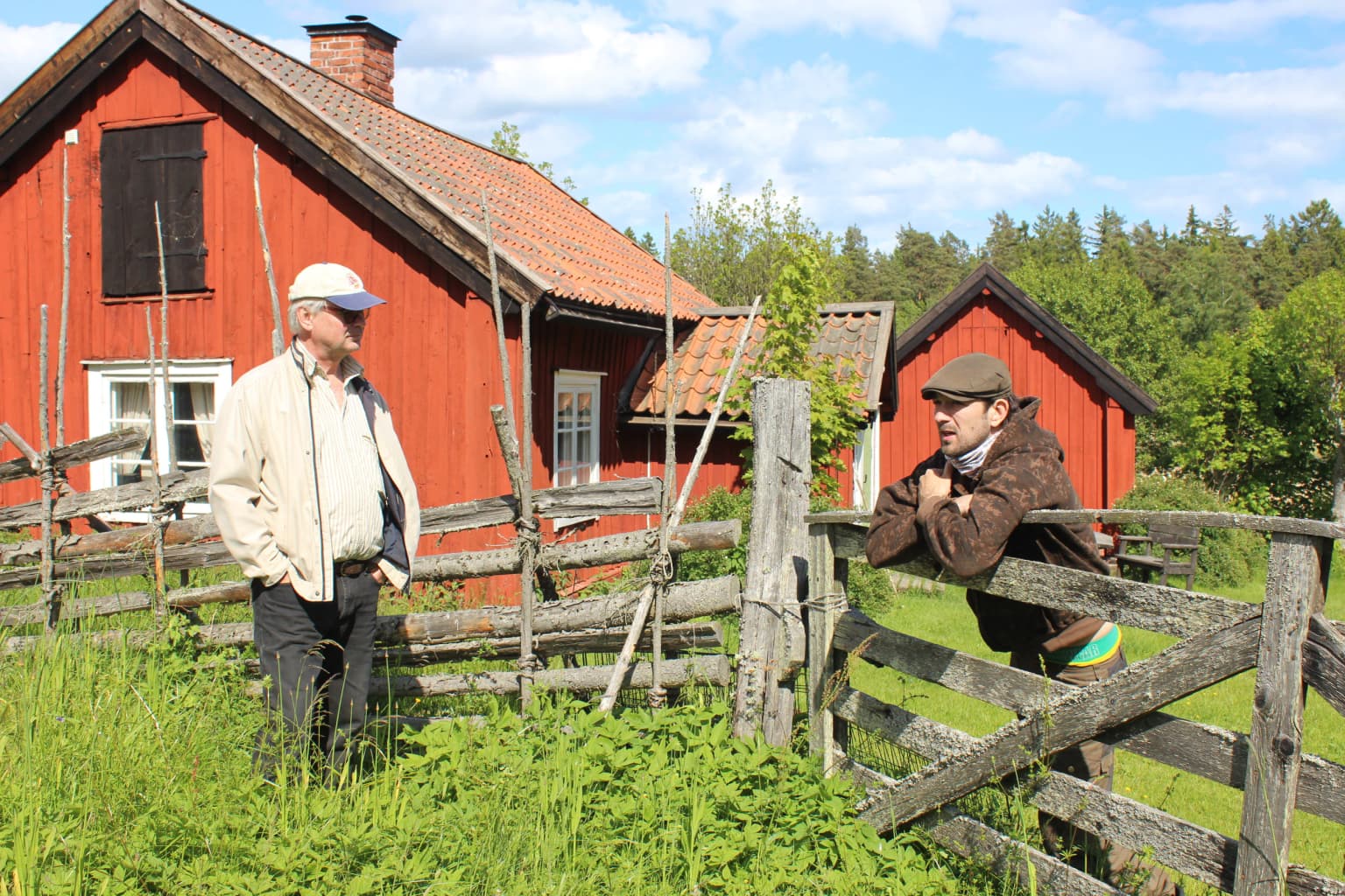 Two individuals standing by a wooden gate in front of red wooden houses with a forested background.