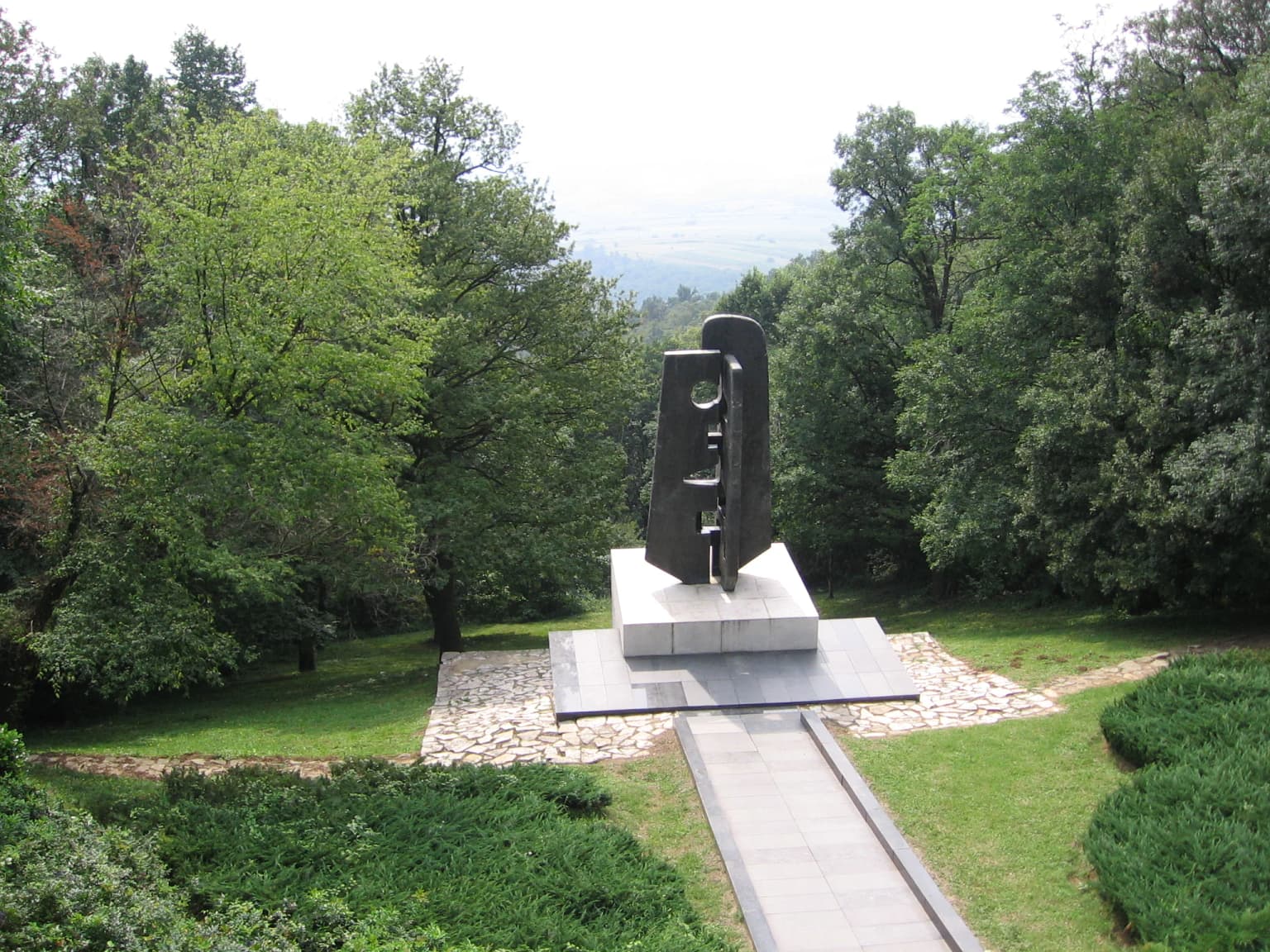 Abstract black monument on stone pedestal with pathway leading to it, surrounded by green trees and grass