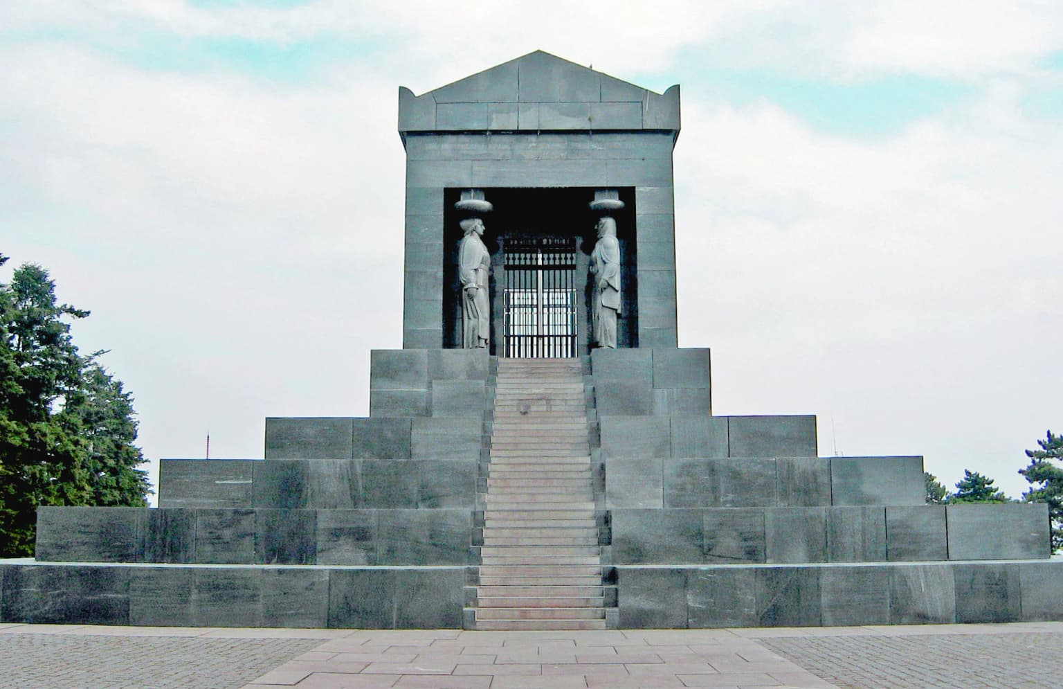 Large stone monument with steps leading up to a structure featuring two statues and a central gate, set against a partly cloudy sky