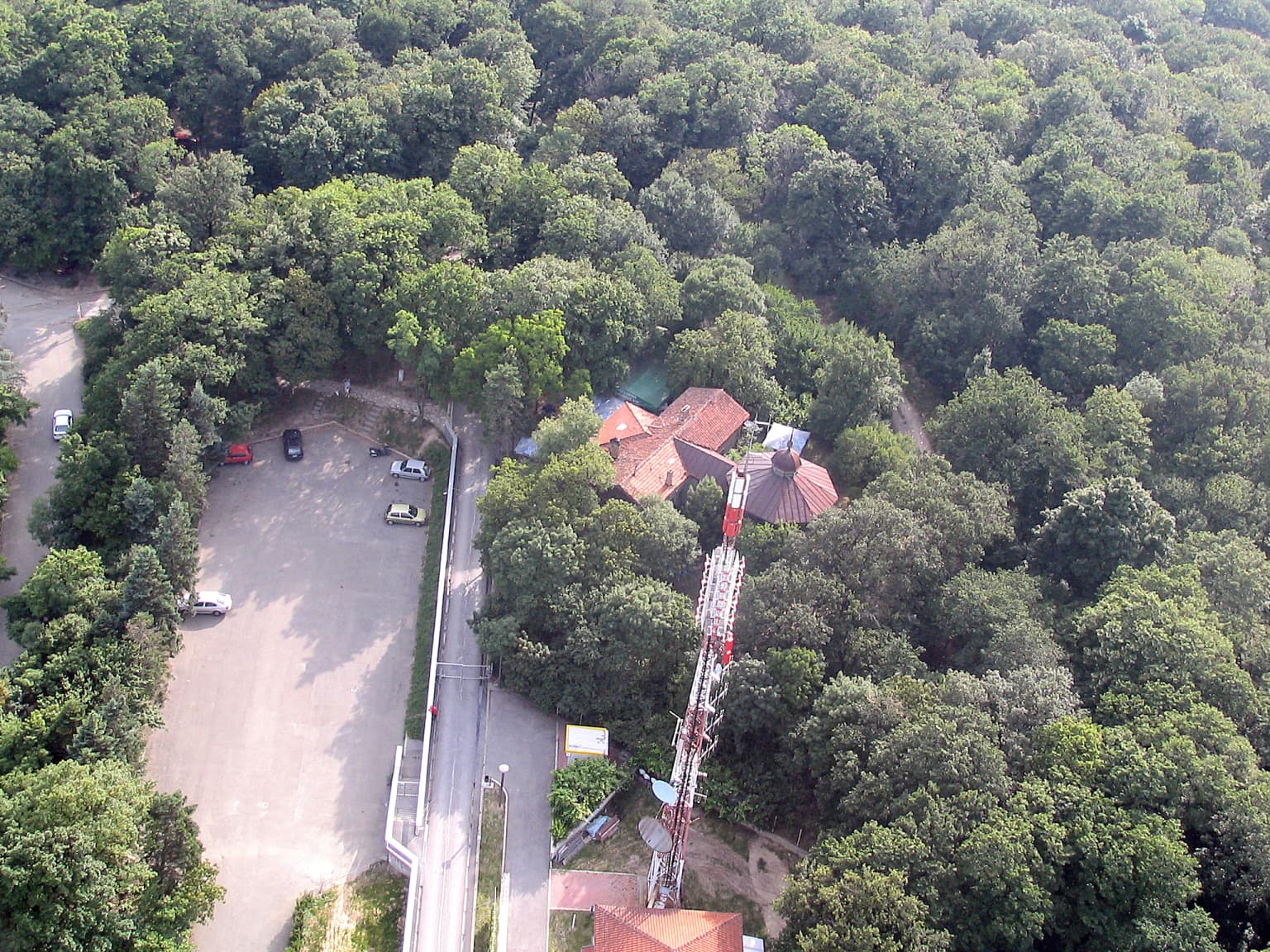 Aerial view showing Mitrovicev dom lodge with red roofs, a tall communication tower, surrounding forest, parking lot, and road