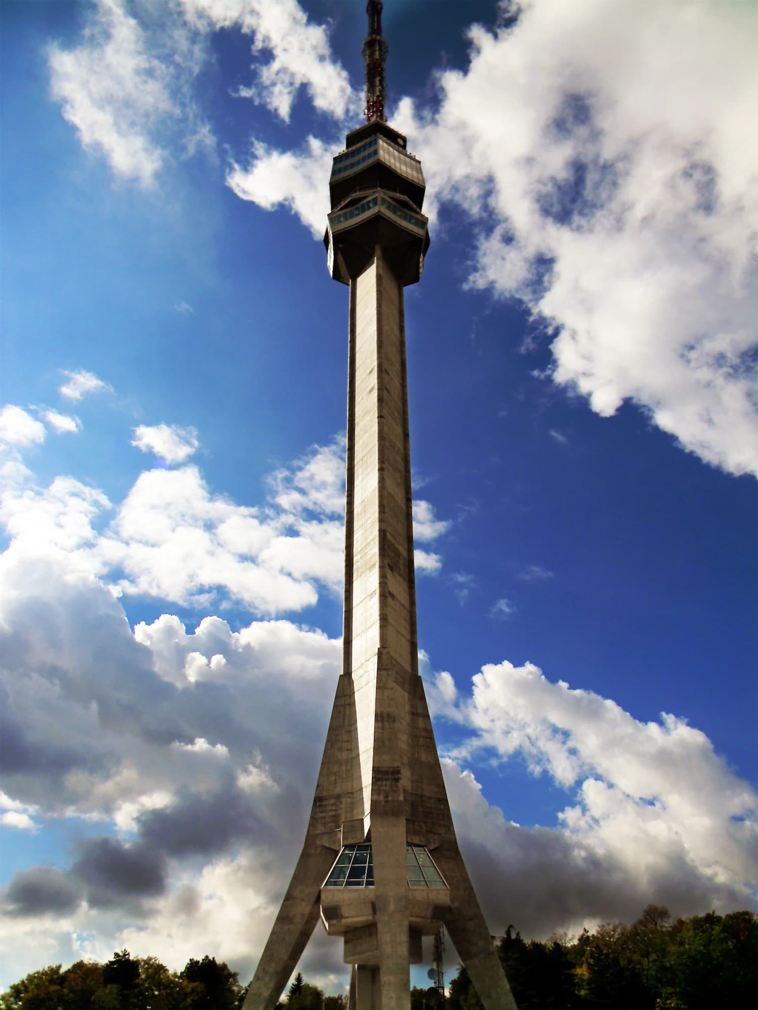Tall concrete tower with three legs against a blue sky with clouds, trees visible at the base.