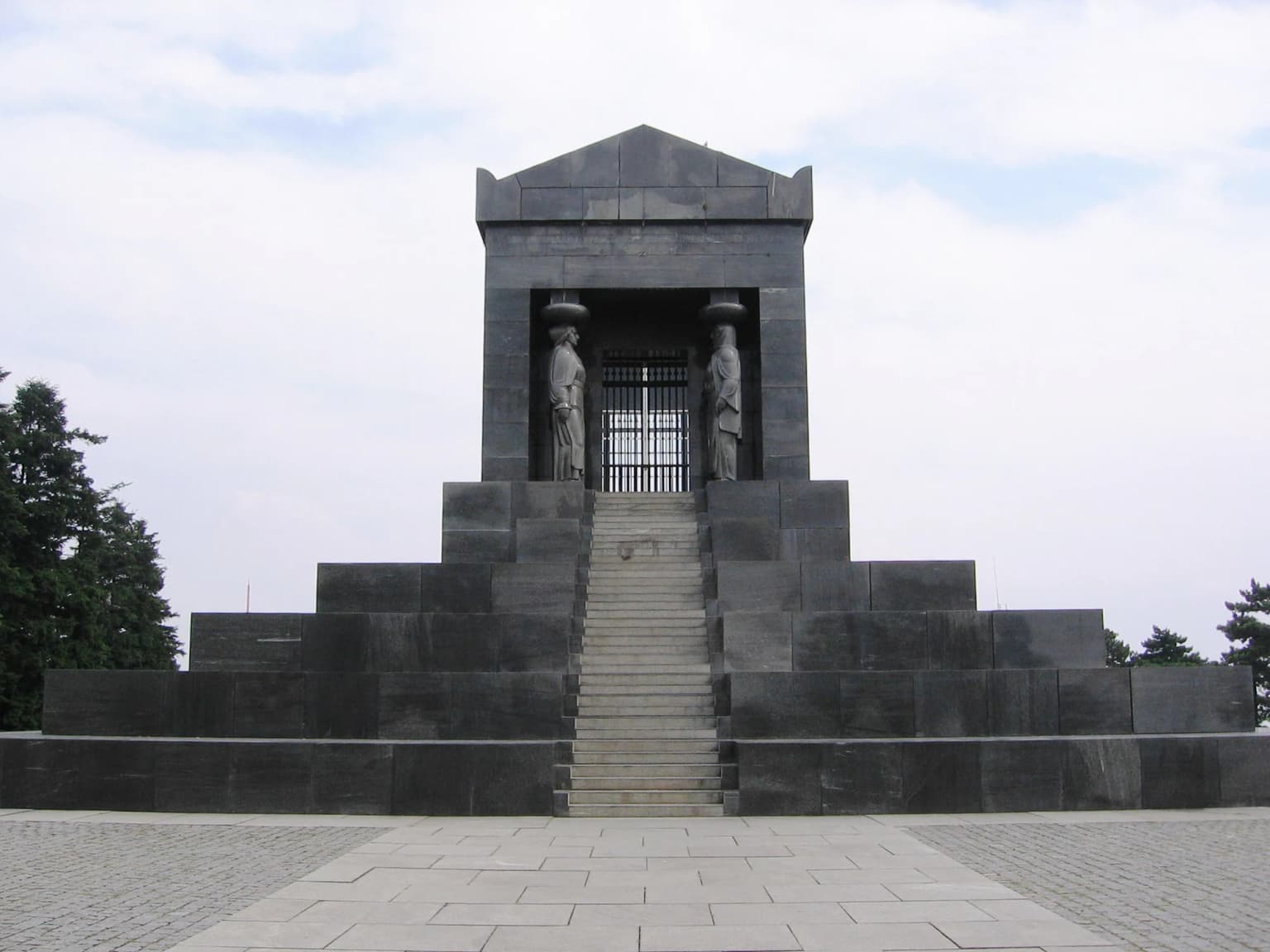 Stone monument with columns and statues, leading up to a staircase on a paved plaza