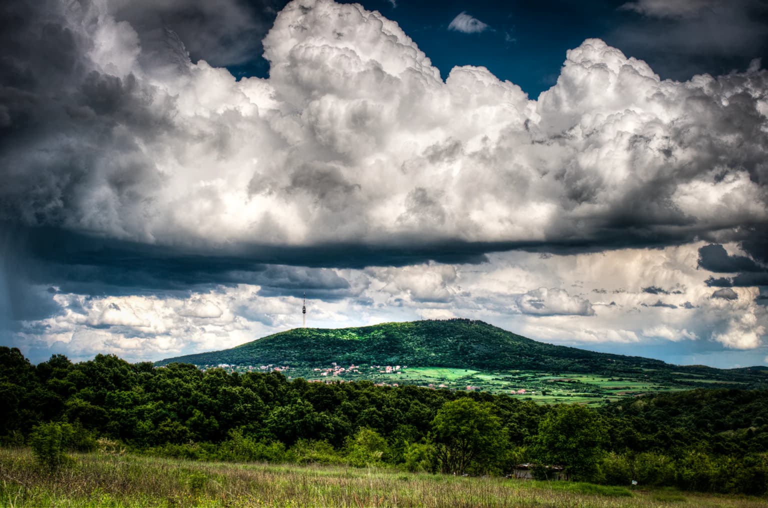 Avala Mountain viewed from a distance with green forests, fields, and large clouds in the sky
