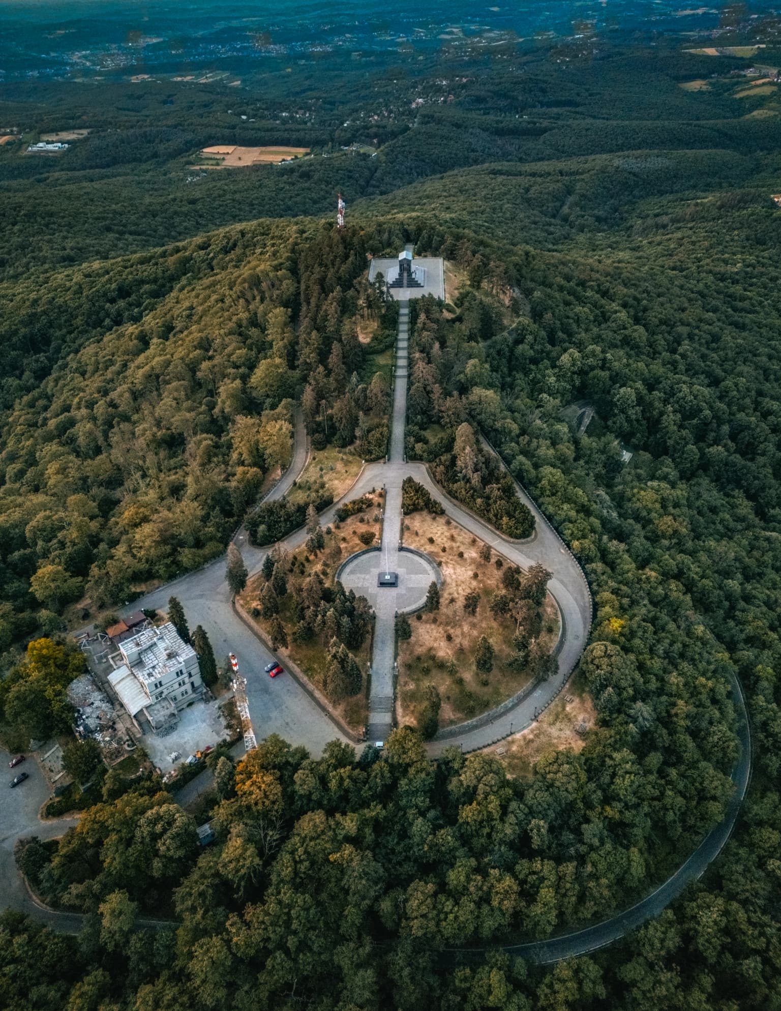Aerial view showing a monument atop a forested mountain with circular roads and pathways leading to the structure