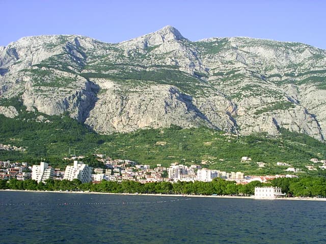 Rocky mountain range with green patches overlooking a coastal town and sea. Buildings line the shoreline with trees between the town and mountains.