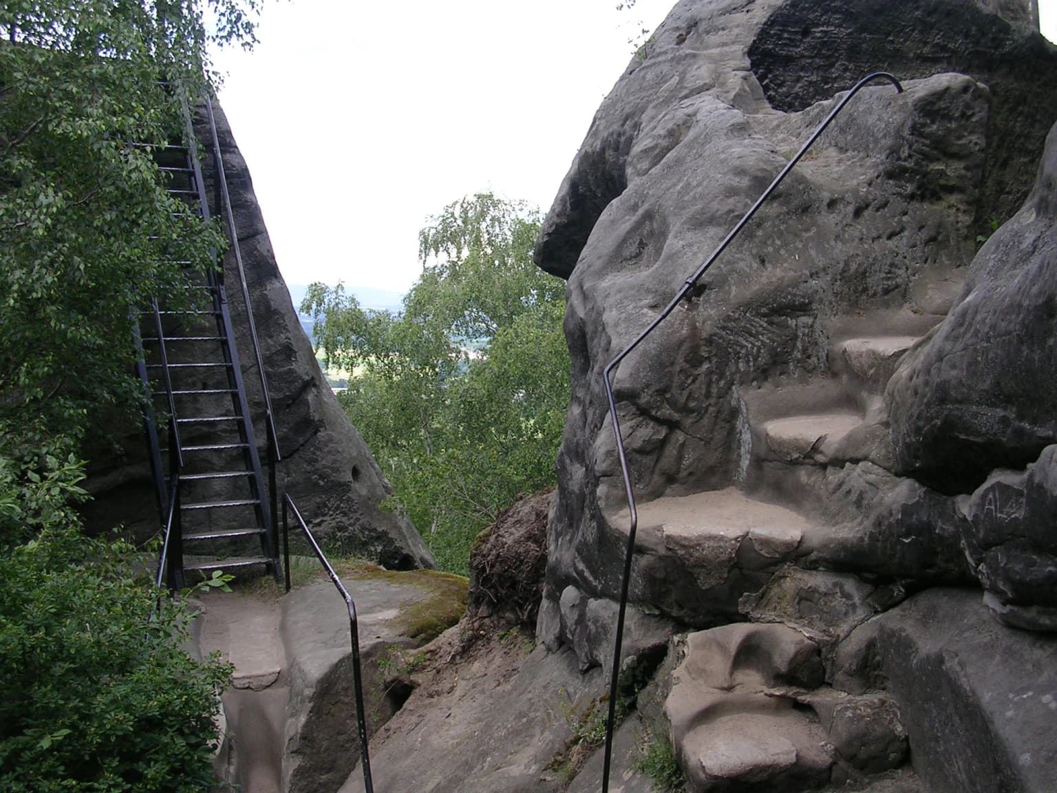 Metal staircase and railings on a rocky cliff face with green vegetation and distant landscape view