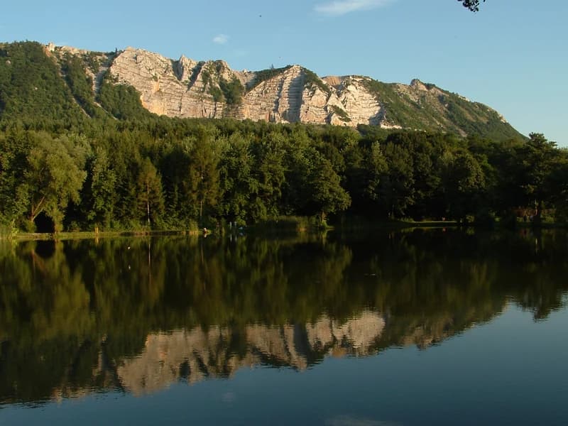 A calm reservoir reflecting a mountain with rocky cliffs and forested slopes under a clear blue sky