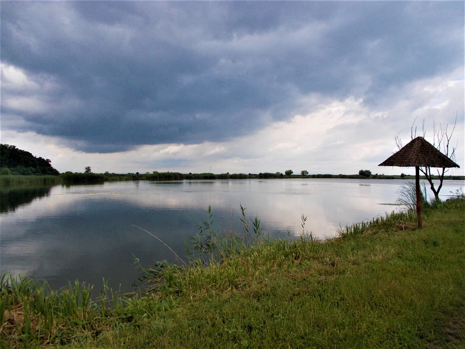 Reflective lake surface, grassy shoreline, wooden shelter, overcast sky with dark clouds