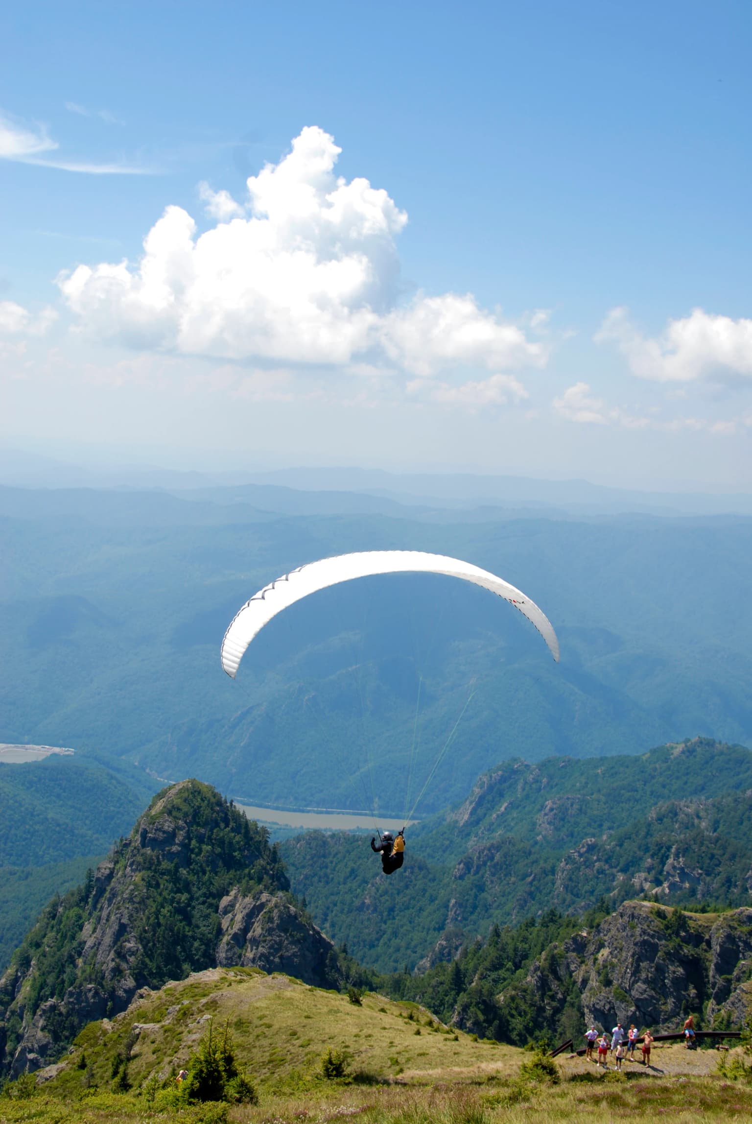 A paraglider in flight over a mountainous landscape with rocky peaks, green valleys, and a blue sky with scattered clouds