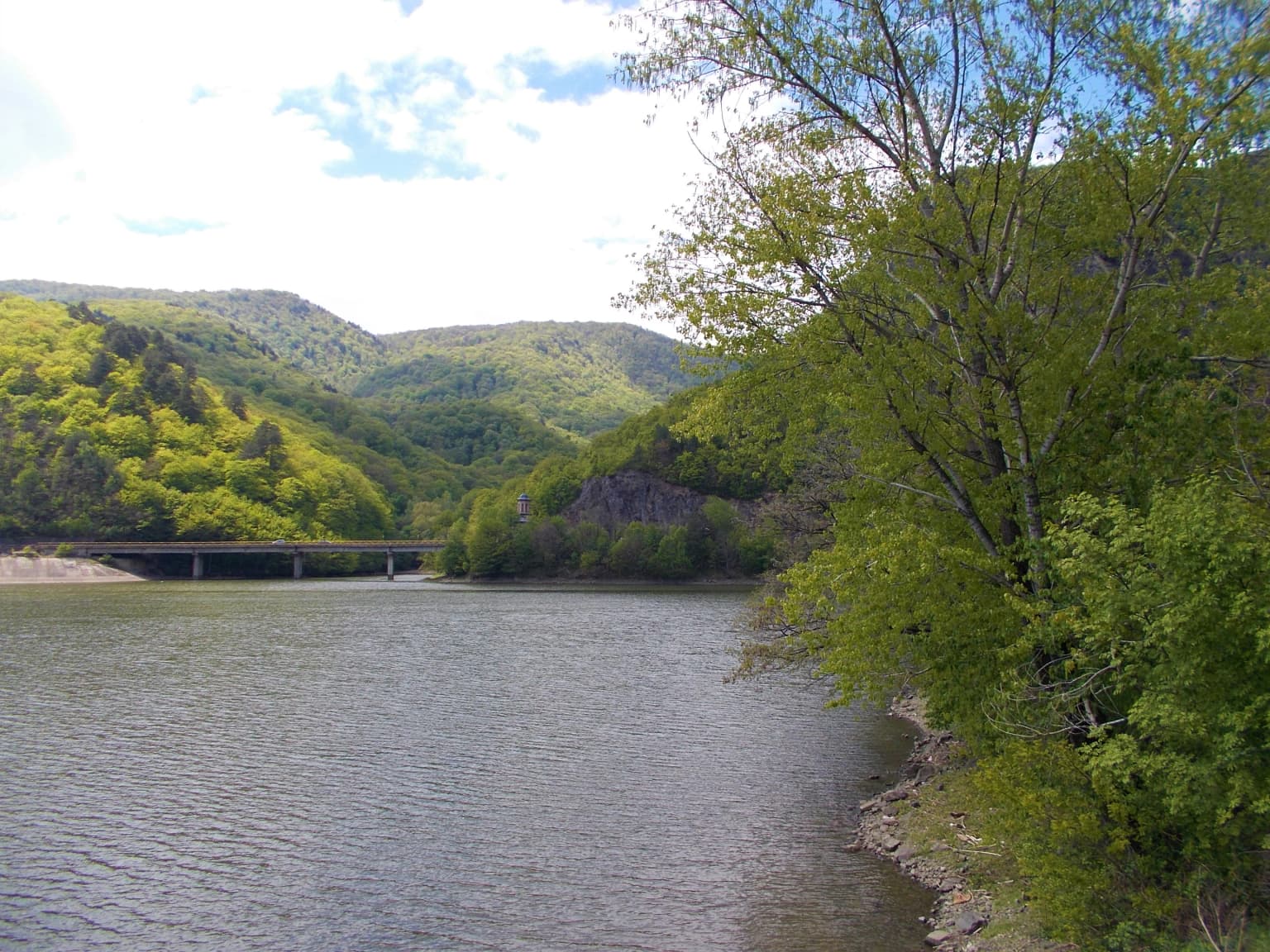 River with bridge spanning across it, surrounded by green hills and trees under a partly cloudy sky