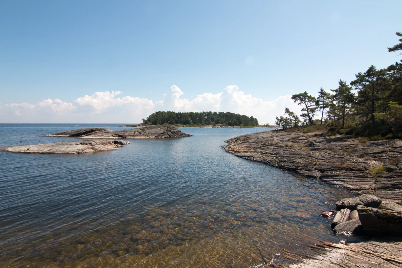 Rocky shoreline with clear water, distant islands, and trees on the right side under a clear blue sky