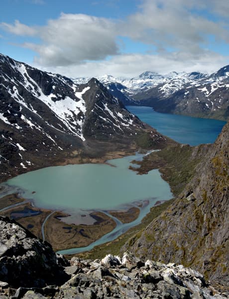 Wide view of mountainous landscape with turquoise lakes, rocky slopes, snow-covered peaks, and a blue sky with clouds