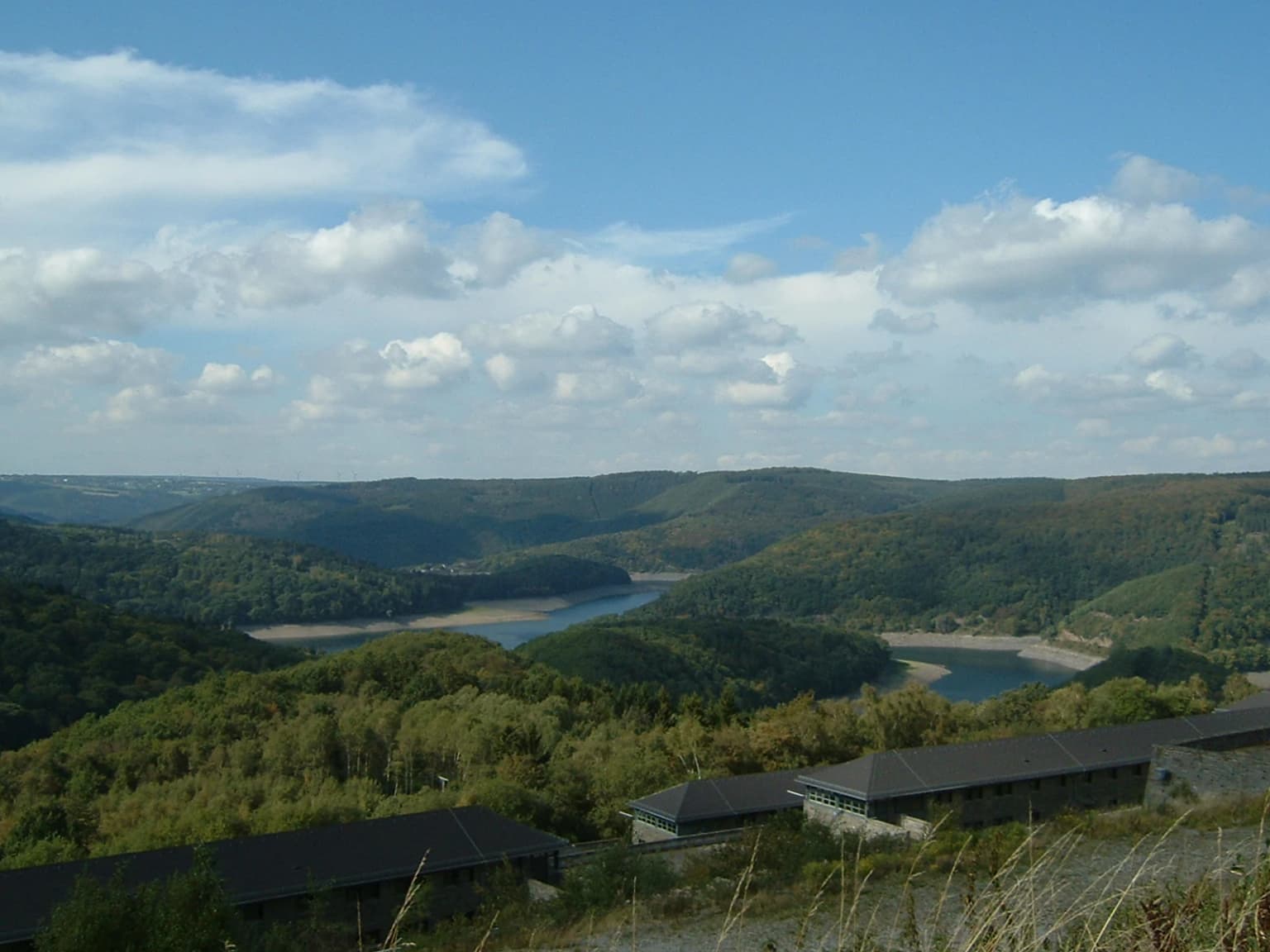 Panoramic landscape view of forested hills, a lake, and distant mountains from Vogelsang Castle with buildings in the foreground.