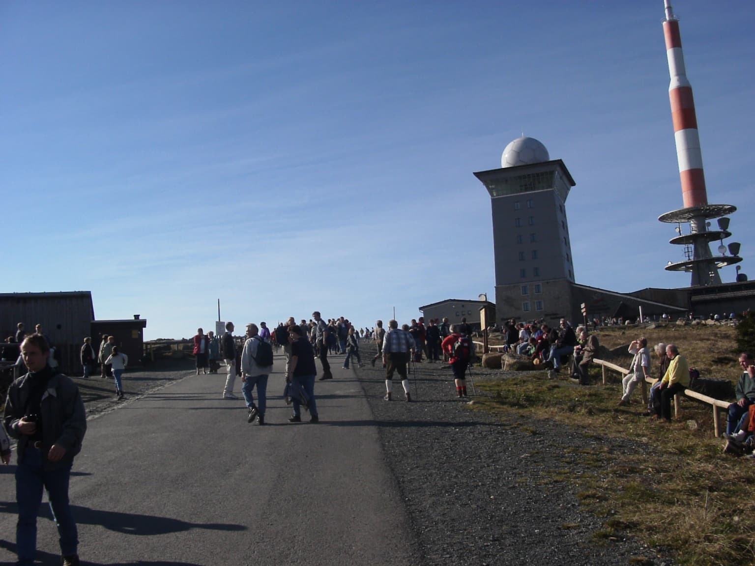 People walking on a paved path at the Brocken summit with an observation tower and radio tower in the background under a clear blue sky
