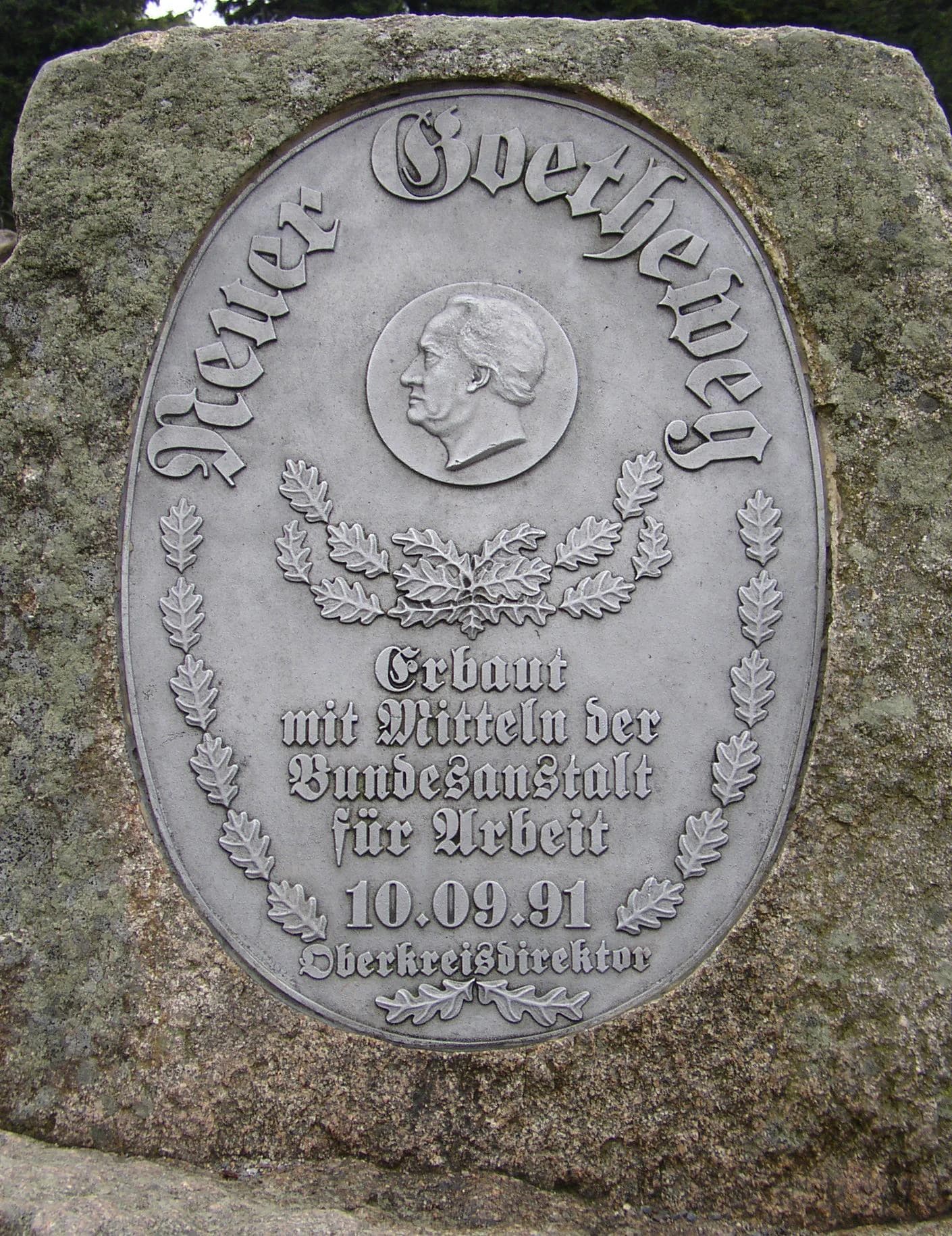 Stone monument with metal plaque showing 'Neuer Goetheweg' text, a profile portrait, oak leaves, and inscribed German text including date 10.09.91