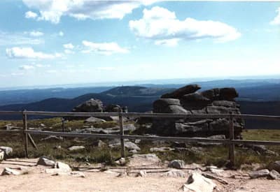 Wide landscape view from Brocken peak featuring large rock formations, metal railing, grassy terrain, and distant mountains under a partly cloudy sky