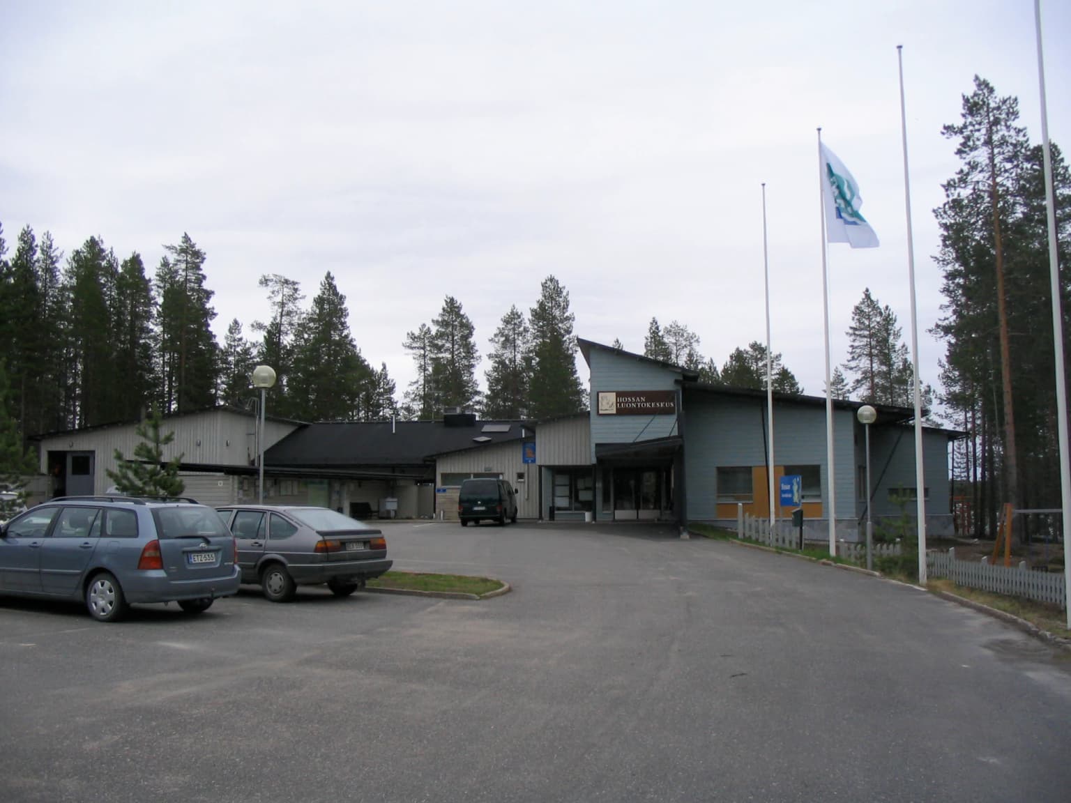 Modern visitor center building with parking lot, cars, and flagpoles surrounded by pine forest
