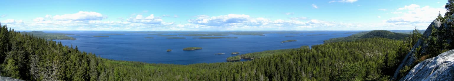 Panoramic view of Lake Pielinen surrounded by forested hills under a partly cloudy sky.