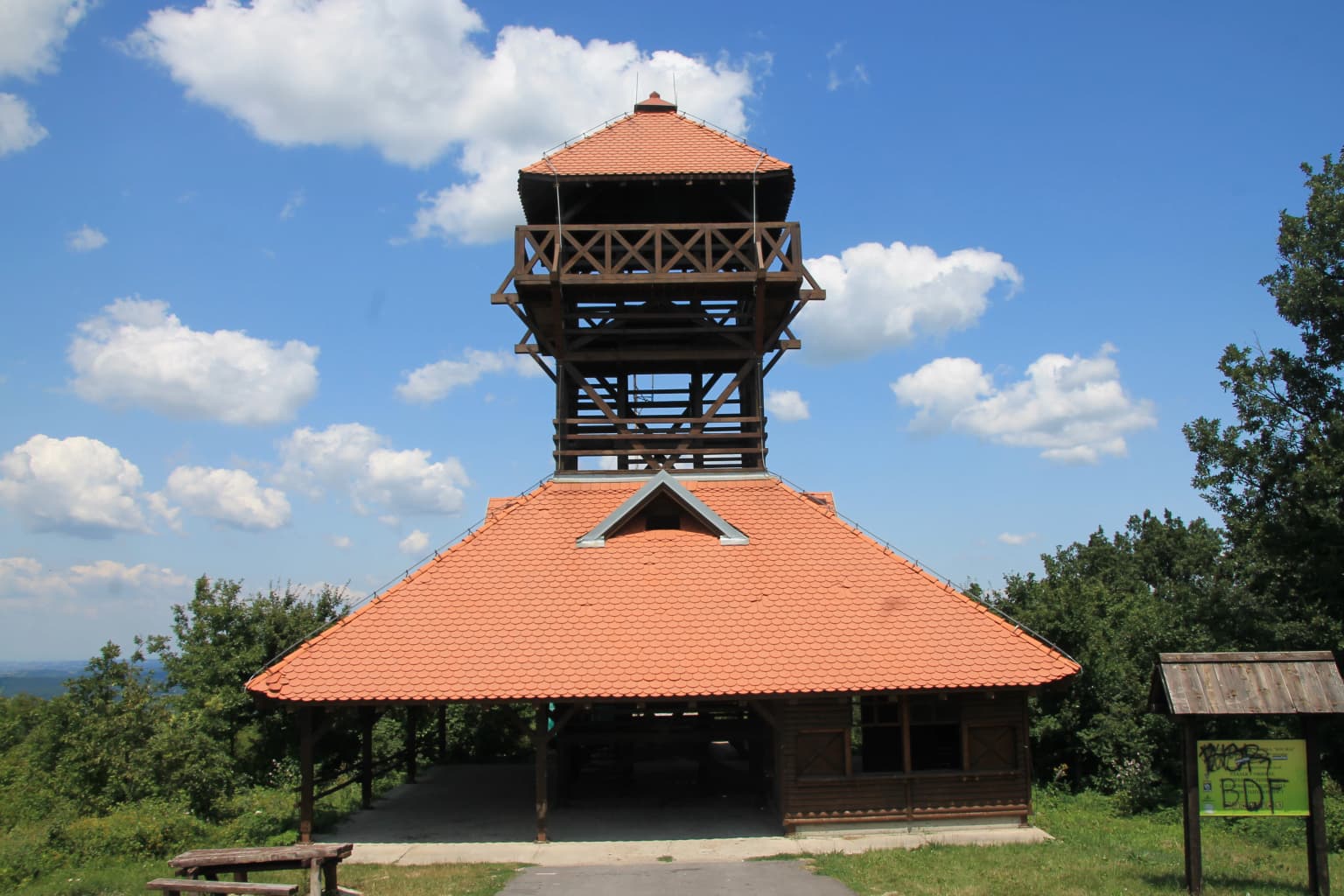 Wooden observation tower with red-tiled roof under blue sky with clouds, surrounded by green trees and a pathway