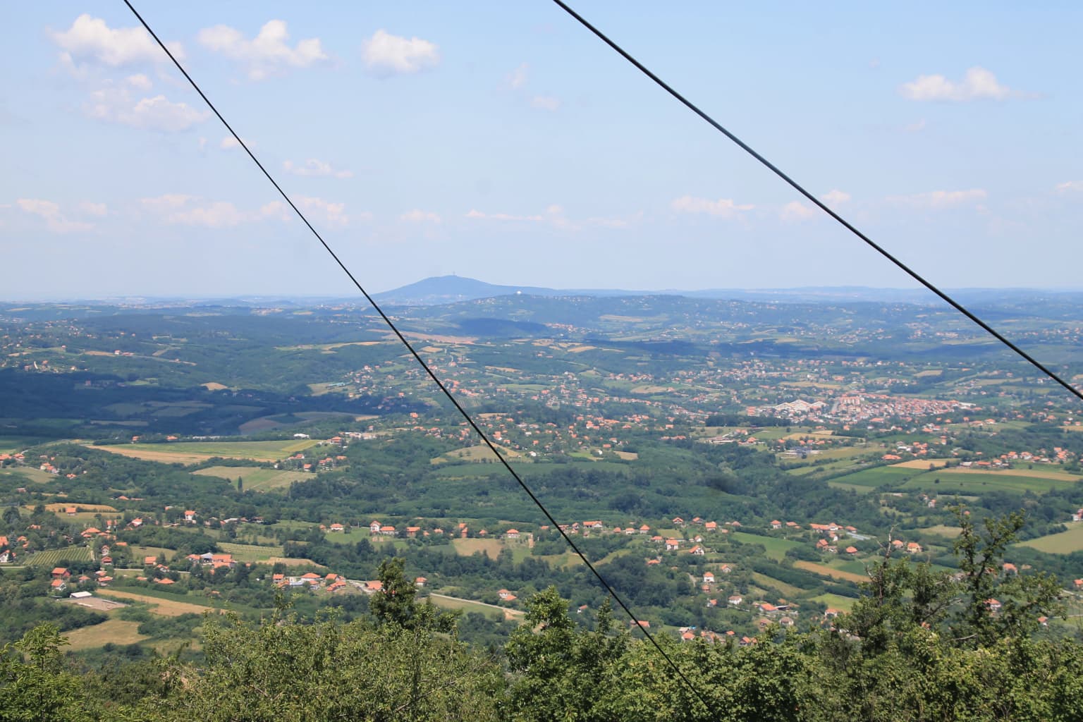 Wide landscape view from a mountain overlooking a valley with fields, scattered houses, and distant hills under a partly cloudy sky