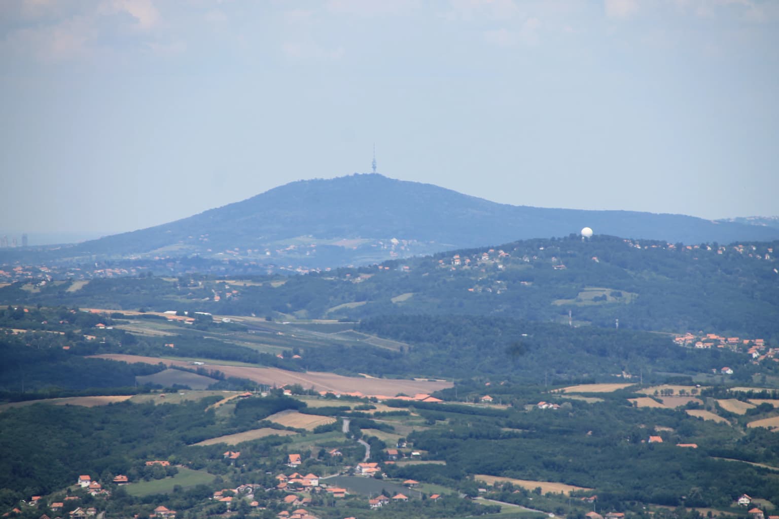 Wide landscape view of Kosmaj mountain showing the mountain peak with a tower, surrounded by rolling hills, agricultural fields, and scattered houses under a partly cloudy sky
