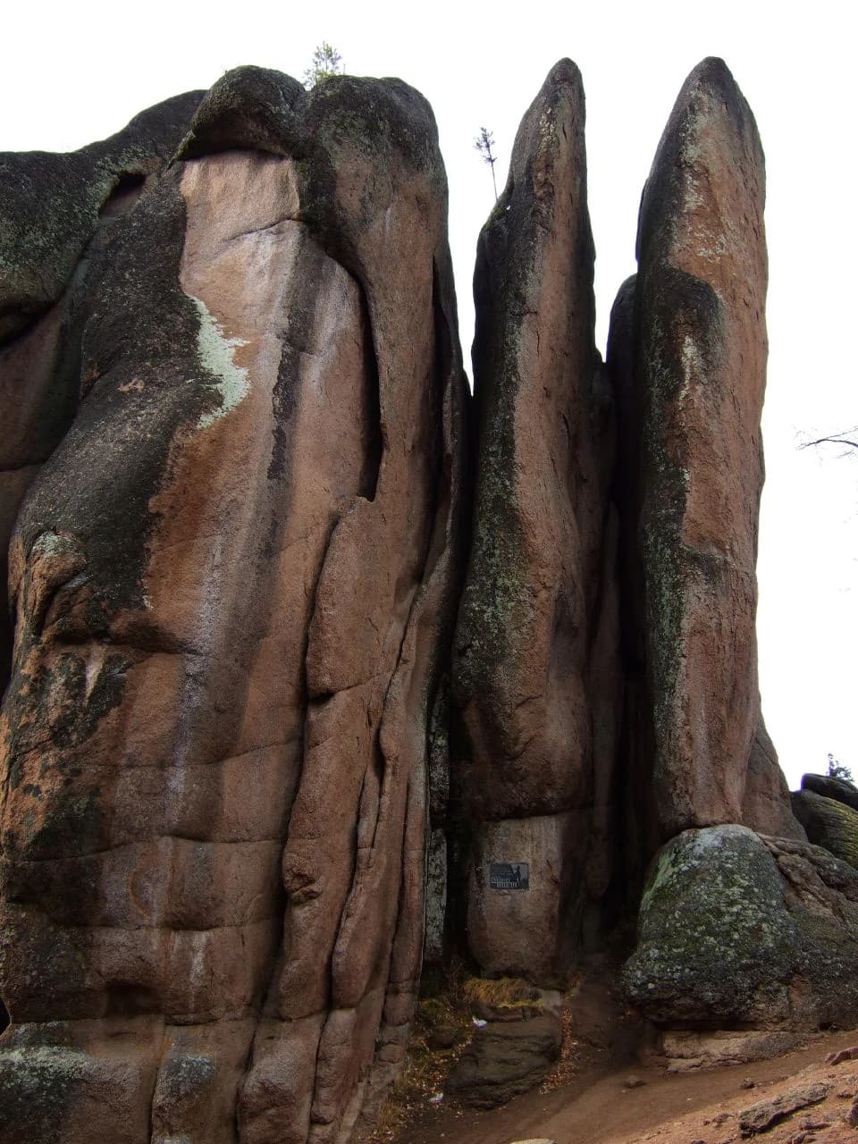 Tall reddish-brown rock pillars with vertical cracks and moss growth, featuring a small blue sign on the central formation