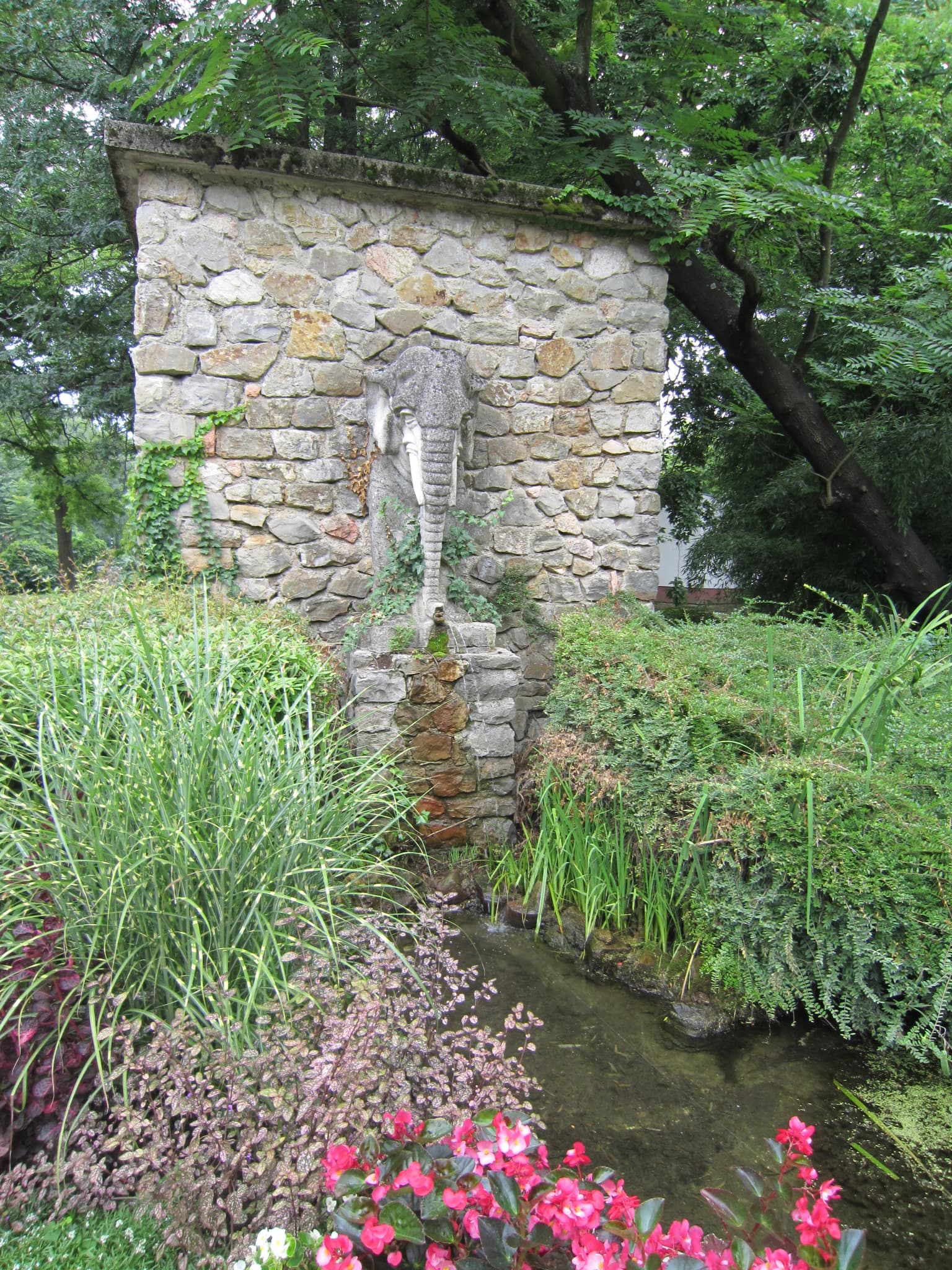 Stone wall with elephant statue fountain, surrounded by green plants and pink flowers with a small stream