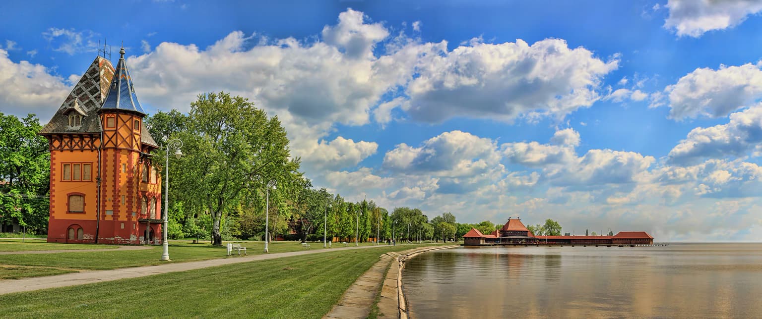 Panoramic view of Lake Palić showing a red-brick building with a steep roof, paved pathway, green grass, trees, and calm lake waters under a partly cloudy sky.
