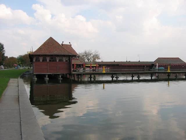 Wooden building on stilts over calm lake water with a long pier and grassy shoreline
