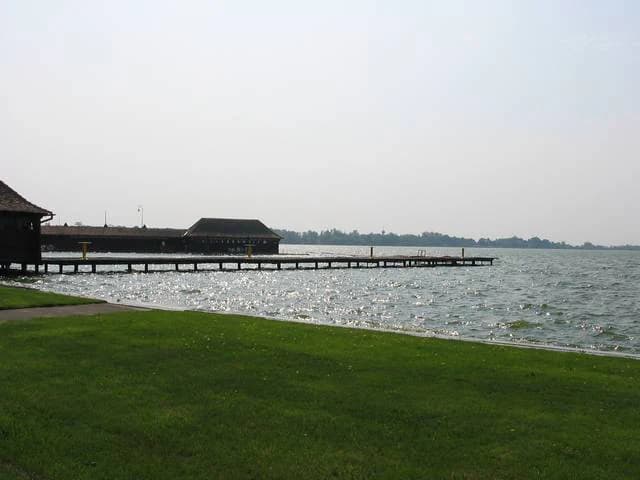 A large body of water with a wooden pier extending from a grassy shore, featuring buildings on the pier under an overcast sky