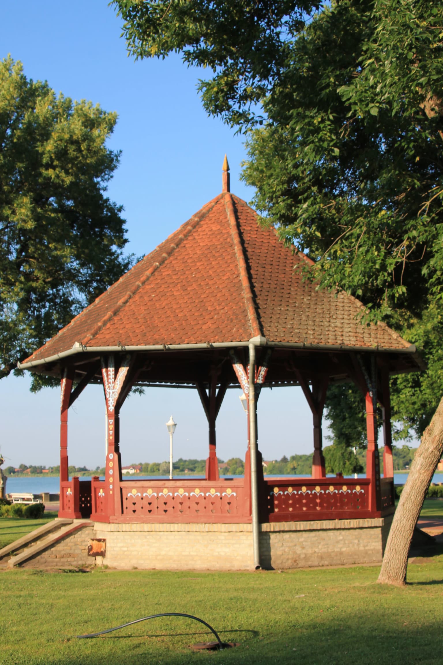 Red-tiled wooden pavilion with decorative railings on a grassy area, surrounded by trees and a body of water in the background