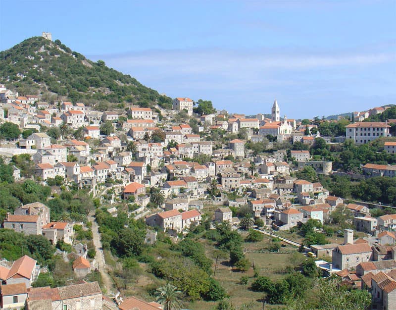 Panoramic view of a coastal town with white buildings and red-tiled roofs on a hillside, featuring a church steeple and a hill in the background