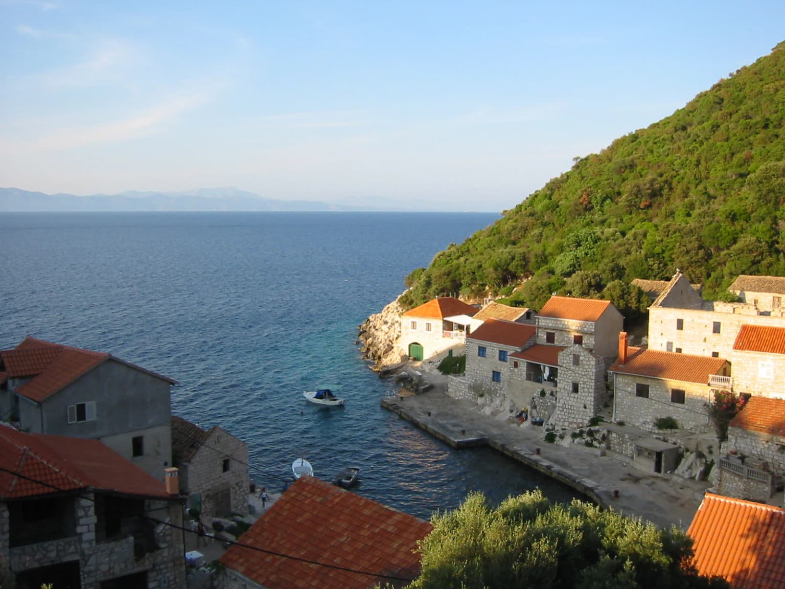 Stone buildings with red roofs lining a calm bay, a small boat anchored near the shore, and a forested hillside in the background