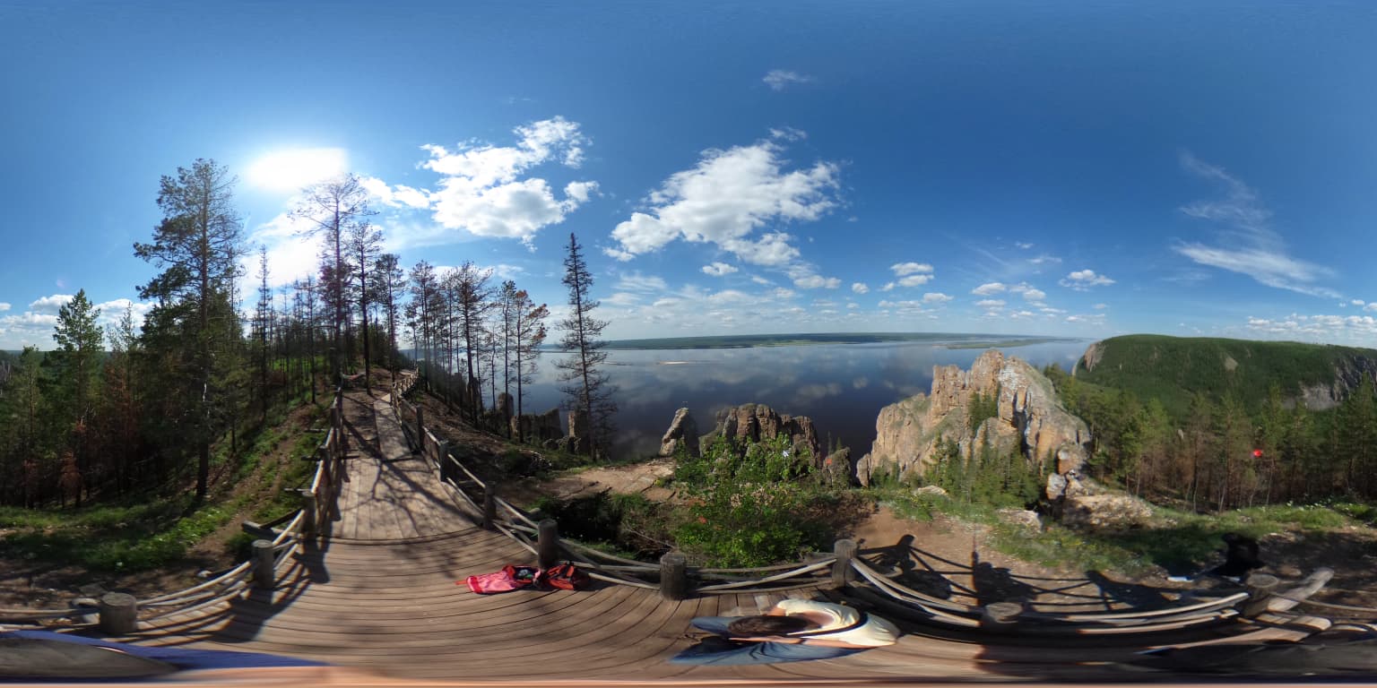 Panoramic view of Lena Pillars rock formations, wooden boardwalk, visitors, trees, and river under a blue sky with scattered clouds.