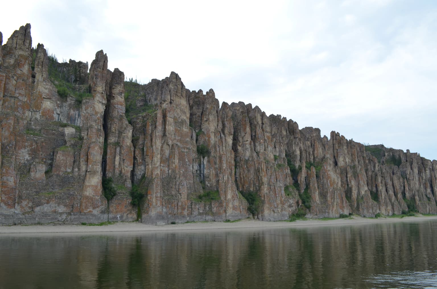 Tall weathered rock pillars rising from a calm river with sandy shore under overcast sky