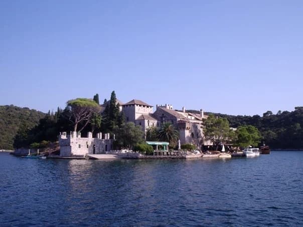 A historic stone building complex on a small island surrounded by calm blue water with trees and hills in the background