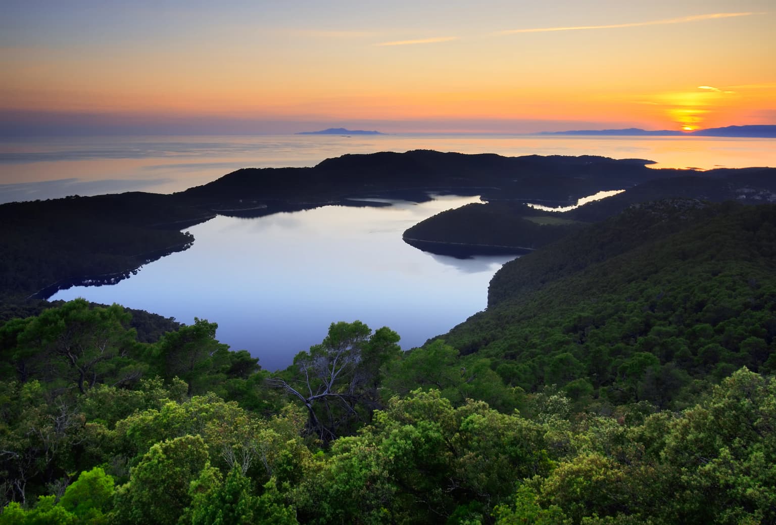 Sunset over a lake surrounded by dense forest with distant hills under an orange and blue sky