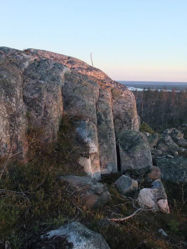 Large rocky cliffs with moss-covered surfaces, surrounded by low vegetation and trees, with a distant view of water under a clear sky