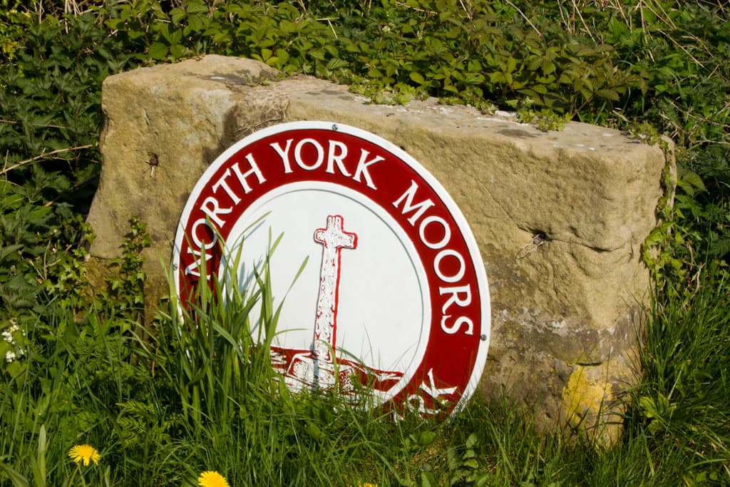Circular sign with 'North York Moors' text, red border, white center with cross and landscape icon, mounted on stone boulder surrounded by green grass and yellow flowers