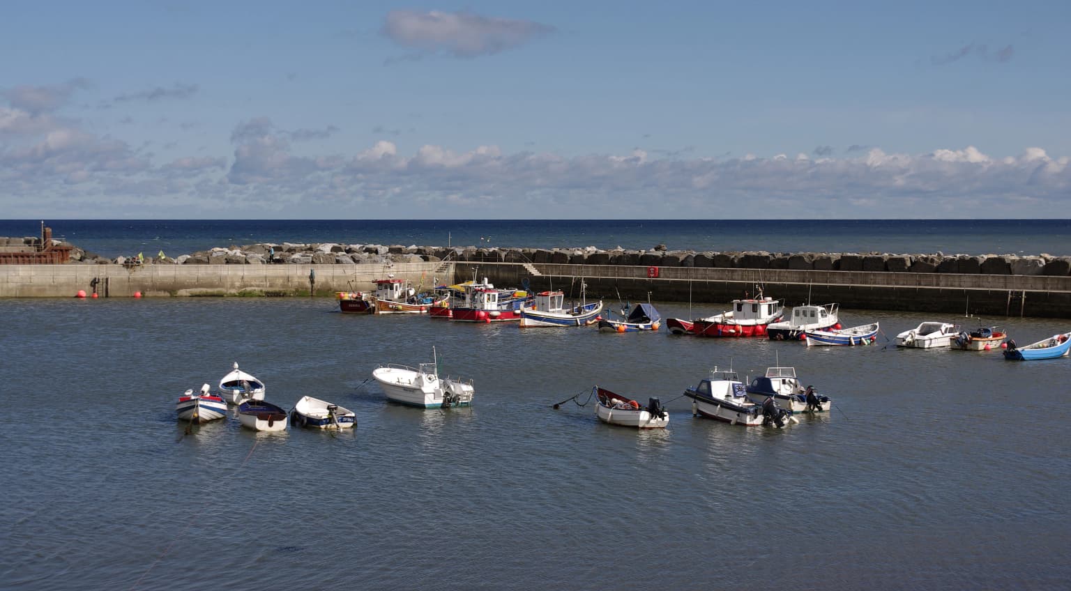 Several small boats floating on calm water in a harbour with stone breakwater and distant sea under a partly cloudy sky