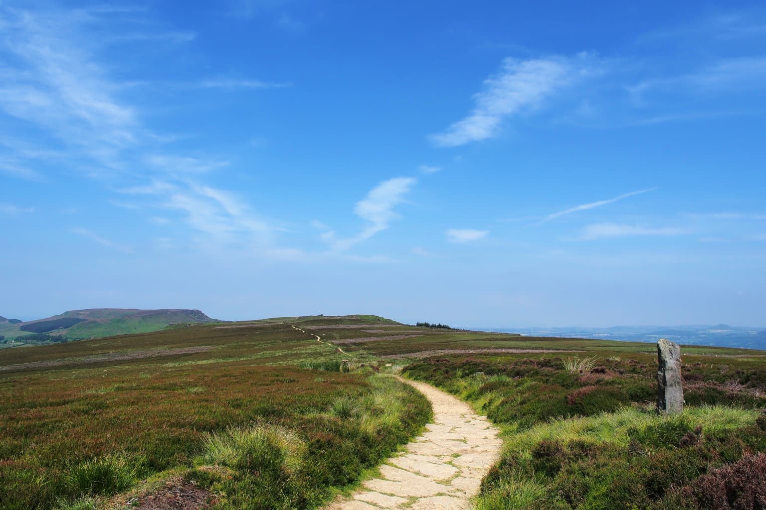 Stone path winding through heather-covered moorland with a stone marker on the right and distant hills under blue sky with wispy clouds