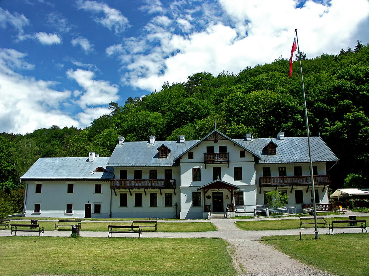 White building with gray roof, flagpole with red flag, grassy lawn, benches, and forested hill