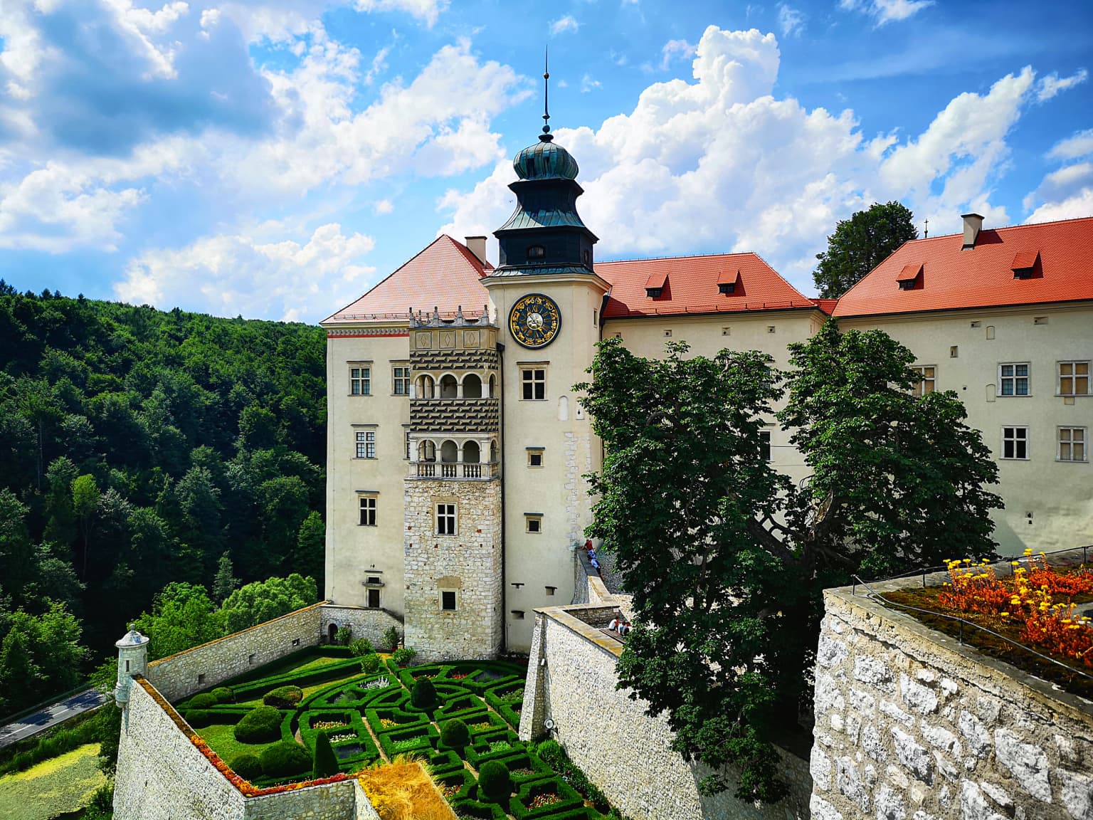 A historic castle with red roofs and a clock tower, surrounded by stone walls and manicured gardens under a partly cloudy sky