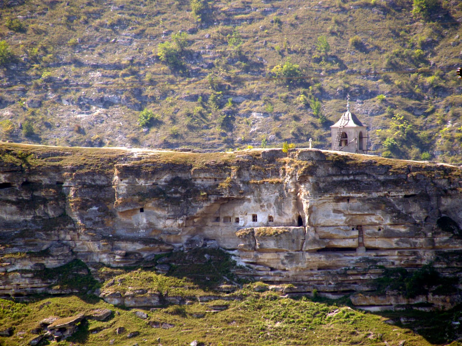 Rocky cliff face with carved cave entrances and a small chapel on the ridge, surrounded by grassy hills