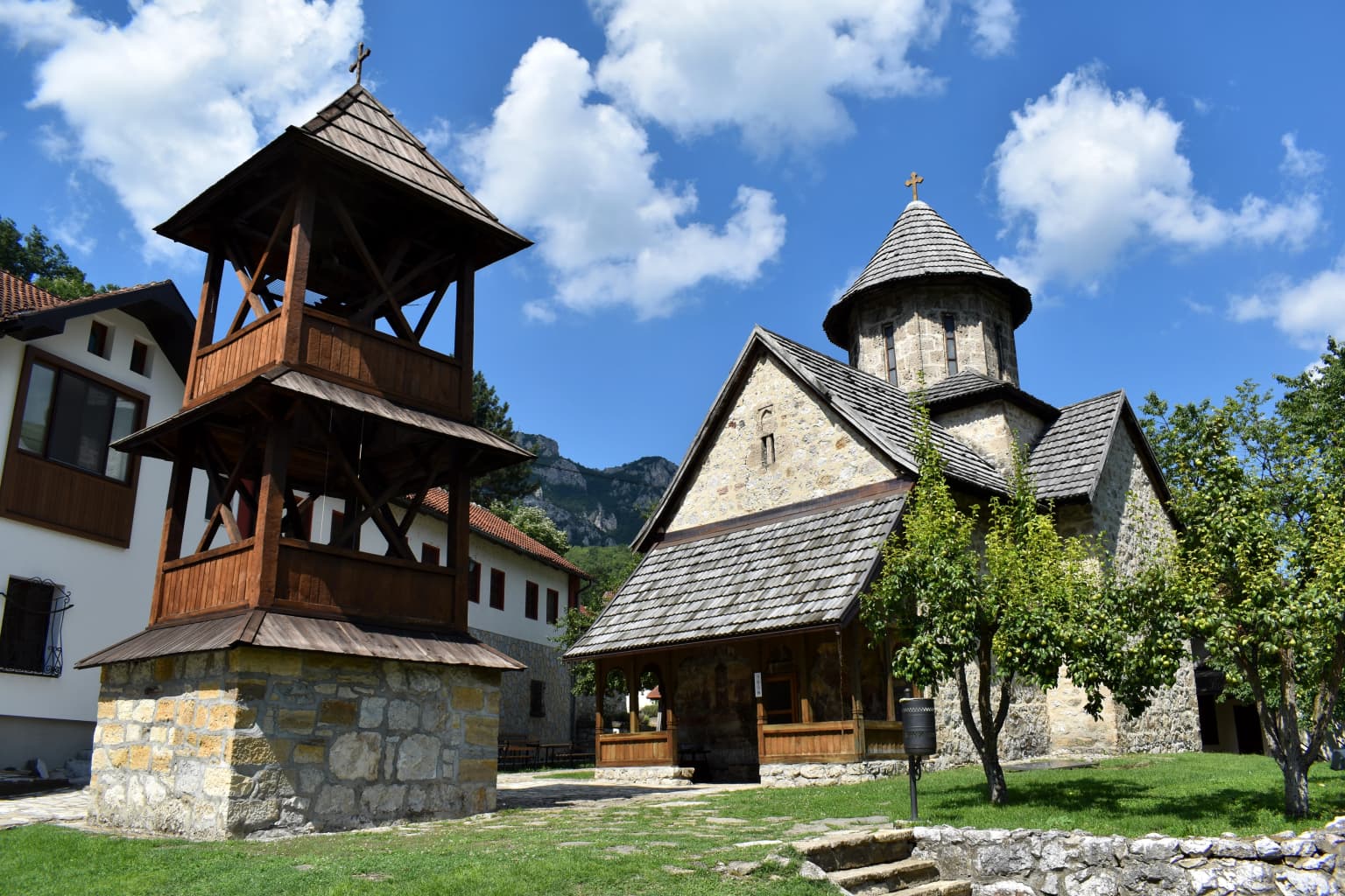 Wooden bell tower with stone base and stone church with conical roof surrounded by green grass and trees under a blue sky with clouds
