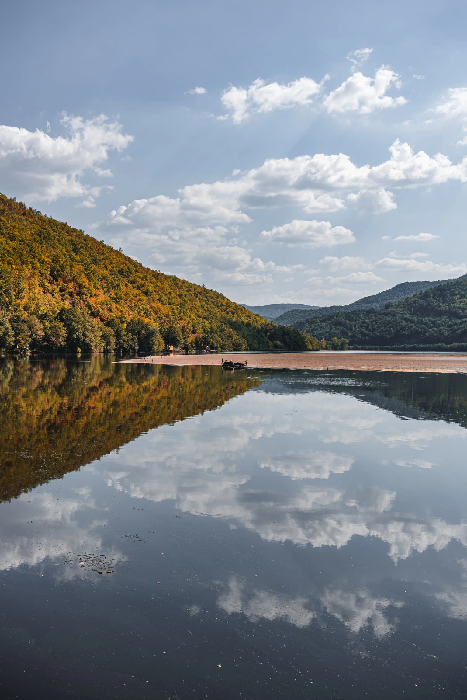 Calm water reflecting forested hills with autumn-colored trees under a partly cloudy sky