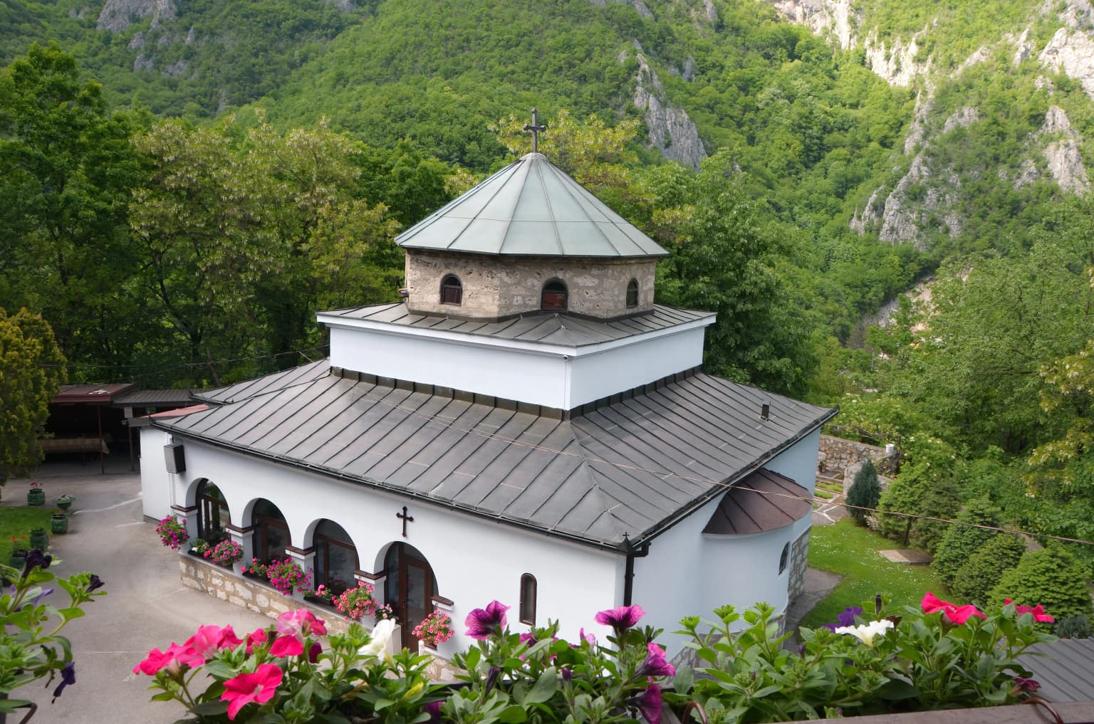 White monastery building with green domed roof, arched windows, and a cross, surrounded by green mountains and pink flowers.